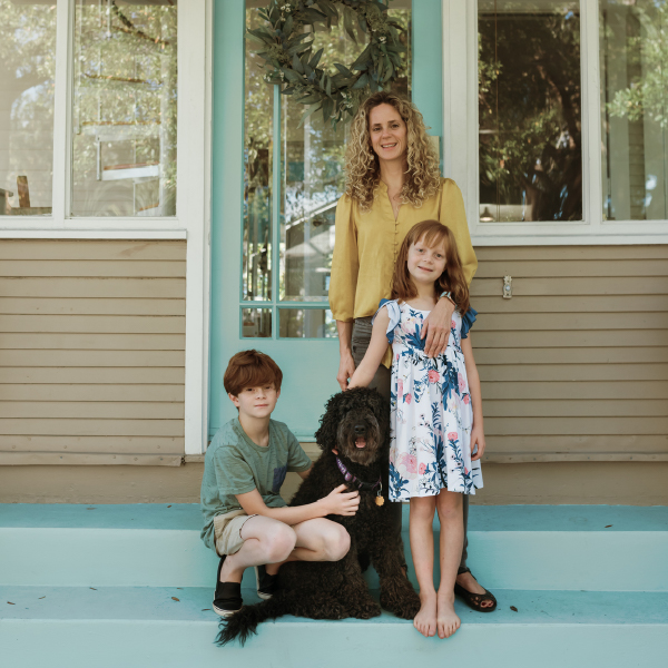Family of three are standing in front of their blue front door with a dark coloured labradoodle. All are smiling at the camera. Pet cremation services.